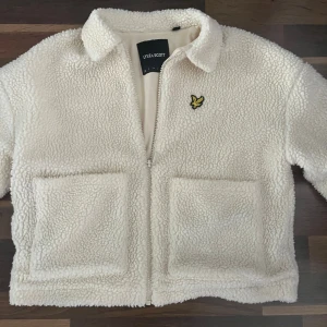 Vit teddyjacka från Lyle & Scott - Mysig vit teddyjacka från Lyle & Scott med gul broderad fågel på bröstet. Jackan har två stora framfickor, krage och dragkedja framtill. Perfekt för dig som gillar chill och stilren look med lite edge.