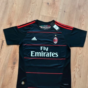 AC Milan Ibrahimović tröja Adidas - Snygg svart AC Milan fotbollströja från Adidas med röda detaljer och klubbmärke på bröstet. Tryck med Ibrahimović och nummer 11 på ryggen. Tillverkad i lätt och ventilerande material med ClimaCool-teknologi. Klassiska röda Adidas-ränder på axlarna.