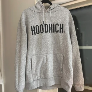 Grå HOODRICH hoodie med tryck - Tja,Säljer en fin grå hoodie från HOODRICH med svart tryck över bröstet pga av ingen användning av den . Tröjan har huva med snörning och en stor magficka framtill. Perfekt för dig som gillar streetwear och vill ha en enkel men snygg look.