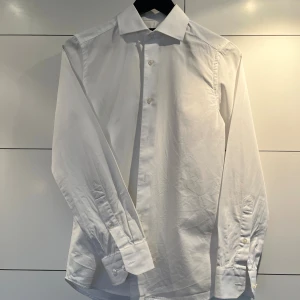 Vit skjorta Riley - Vit skjorta från Riley i super slimfit-modell med klassisk krage och knappar framtill. Skjortan har långa ärmar med manschett och är gjord i mjuk bomull. Perfekt för dig som gillar en stilren och tajt passform.