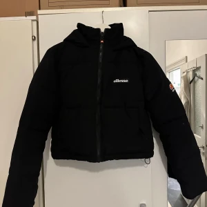 Svart pufferjacka från Ellesse - Svart pufferjacka från Ellesse med huva och dragkedja framtill. Jackan är croppad och har Ellesse-logga på bröstet och ryggen. Perfekt för kalla dagar och har en sportig vibe. Tillverkad i syntetmaterial och har långa ärmar.