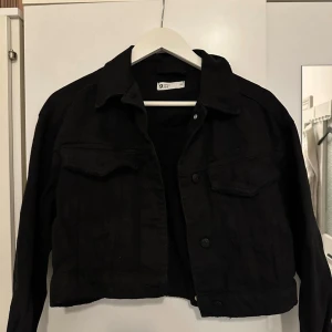 Svart croppad jeansjacka från Gina Tricot - Svart croppad jeansjacka från Gina Tricot med klassisk krage, knappar framtill och två bröstfickor. Jackan har långa ärmar och en snygg, rak passform som ger en cool vibe till outfiten.