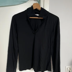 Svart långärmad tröja med krage - Svart långärmad tröja med snygg krage och v-ringad öppning. Tröjan är i mjukt bomullsmaterial och har en clean, minimalistisk look som passar perfekt till jeans eller kjol. Enkel att styla och superbekväm för alla tillfällen.