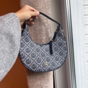 Blå axelväska med monogram - Snygg axelväska blått grått med svart monogrammönster över hela väskan. Väskan har en halvmåneform, guldfärgade detaljer och dragkedja upptill. 