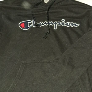 Svart Champion hoodie med broderad logo - Svart hoodie från Champion i storlek L med broderad logga i vitt, blått och rött på bröstet. Klassisk känguruficka framtill och justerbar huva med snörning. Perfekt för en chill och sportig stil.