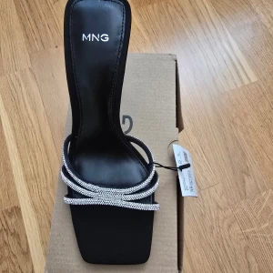 Svarta sandaler med strass från MNG - Snygga svarta sandaler från MNG med fyrkantig tå och smala remmar täckta av glittrande strass. Skon har hög klack och en elegant design som verkligen sticker ut. Perfekta för dig som vill ha något extra till outfiten.