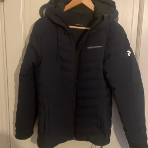 Svart pufferjacka Peak Performance S - Snygg svart pufferjacka från Peak Performance med huva och diskret logga på bröstet och ärmen. Jackan har dragkedja framtill och två fickor. Perfekt för kalla dagar och har en stilren, sportig look.