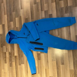 Blå mjukisbyxor från Nike, storlek L (147-158 cm) och blå Nike tech fleece, storlek XL (158-170 cm)  - Säljer ett par blå mjukisbyxor från Nike med svart logga på benet.Och en Nike fleece med svart logga på bröstet. Byxorna har resår i midjan och vid bensluten för skön passform. Materialet är mjukt och passar perfekt för chill eller träning. Klassisk sportig look med enkel design.
