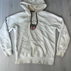 Vit GANT hoodie  - Tjena säljer nu en skön Vit hoodie från GANT med broderad logga och emblem på bröstet. Tröjan har huva med svarta snören och en stor magficka framtill. Mjuk bomullskvalitet och ribbade muddar vid ärmslut och nederkant. Perfekt för chill dagar.