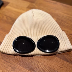 Beige Goggle-mössa från C.P. Company - Ribbstickad beige mössa från C.P. Company med ikoniska svarta goggle-linser framtill. Mössan har uppvikt kant och är tillverkad i mjuk bomull som ger en skön passform. Perfekt för dig som vill sticka ut med en unik streetwear-accessoar. Den mössan är lite smutsig på baksidan 