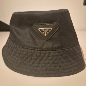 Svart bucket hat från Prada - Stilren svart bucket hat från Prada med klassisk trekantig logotyp framtill. Hatten har rundad form och är tillverkad i ett slätt syntetmaterial som ger en modern vibe. Perfekt accessoar för att lyfta din streetwear-look.