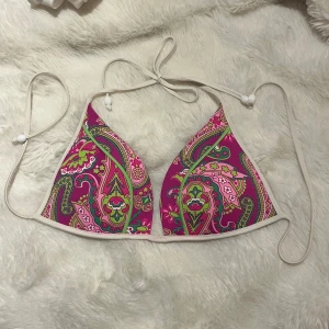 Färgglad bikiniöverdel från Calzedonia - Snygg bikiniöverdel från Calzedonia med färgstarkt paisleymönster i rosa, grönt och inslag av gult. Triangelformad modell med vita knytband och små pärldetaljer. Perfekt för strandhäng och sommarvibbar!