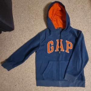 Blå fleece hoodie från GAP Kids - Blå hoodie från GAP Kids med orange foder i huvan och stora orange bokstäver på bröstet. Tröjan har dragkedja framtill, huva och praktisk ficka. Tillverkad i mjuk fleece som är perfekt för kyliga dagar. Snygg och bekväm för en avslappnad stil.