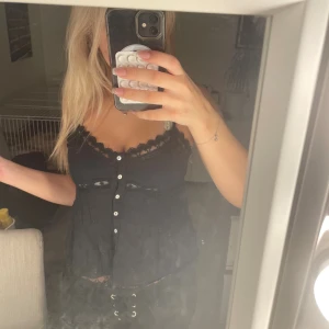 Zara Linne - Säljer en svart linnetopp med spetskant och dekorativa vita knappar framtill. Toppen har v-ringad hals och fina spetsdetaljer både vid halsen och under bysten 