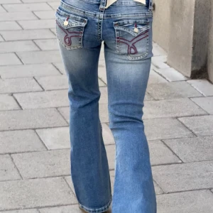 Blå bootcut jeans med broderade fickor - Snygga blå bootcut jeans med låg midja och slitningar på knäet. Baktill har de två fickor med röd broderad detalj och knappstängning. Jeansen har en klassisk tvättad look och är tillverkade i jeansmaterial. Perfekt för dig som gillar retrovibbar och coola detaljer.