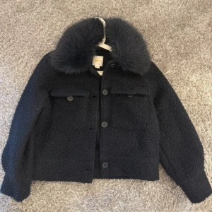 Mörkblå teddyjacka med fuskpäls-krage - Säljer en mörkblå teddyjacka med stor fluffig fuskpäls-krage. Jackan har två bröstfickor med lock och knappar framtill. Storlek 158/164 men passar XS/S