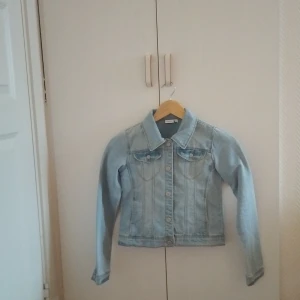 Ljusblå jeansjacka från Name It -  Ljusblå jeansjacka från Name It med klassisk krage, bröstfickor med knappar och markerade sömmar. Jackan har långa ärmar och stängs med metallknappar framtill. Endast använd några få gånger.