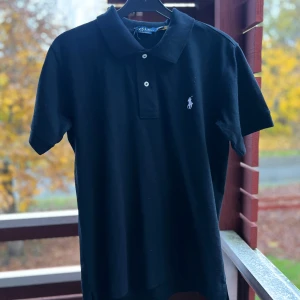 Svart piké från Polo Ralph Lauren - Svart pikétröja från Polo Ralph Lauren med klassisk krage och två vita knappar framtill. Slim fit-modell med korta ärmar och den ikoniska broderade loggan på bröstet. Tillverkad i mjuk bomull som är skön mot huden. Perfekt för en clean och stilren look.