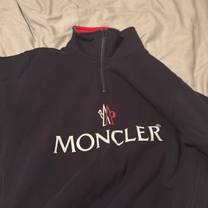 Svart Moncler half zip sweatshirt - Svart sweatshirt från Moncler med stor vit logotyp och röd detalj framtill. Tröjan har en half zip dragkedja vid halsen och insida i rött. Långärmad och tillverkad i mjukt material som känns skönt mot huden. Perfekt för dig som gillar exklusiva märken.