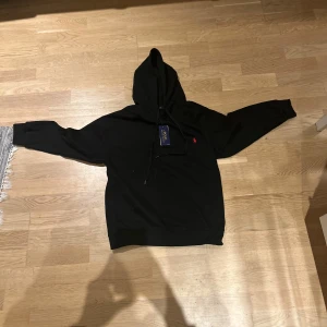 Svart hoodie från Polo Ralph Lauren - Svart hoodie från Polo Ralph Lauren med klassisk huva och snörning. Tröjan har en röd broderad logga på bröstet och en stor magficka framtill. Tillverkad i mjuk bomull och har långa ärmar. Perfekt för chill dagar och streetwear vibes. Helt ny lappen fortfarande på endast testad en gång men den var för liten.