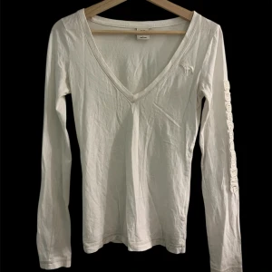 Vit långärmad topp Abercrombie - Vit långärmad topp från Abercrombie & Fitch med djup v-ringning och broderad logga på bröstet. Ärmen har texten 'Abercrombie' broderad längs sidan. Perfekt basic med snygg passform.