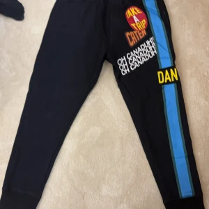 DSQUARED2 byxor - Svarta mjukisbyxor från DSQUARED2 med coola tryck och färgglada detaljer. Byxorna har text och grafik i gult, rött, blått och grönt, samt blå ränder längs benet och röda lönnlöv. Tillverkade i mjukt bomullsmaterial och har en relaxed passform med muddar vid benslut. Äkta!