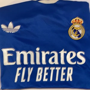 Blå Real Madrid tredjetröja 25/26 - Snygg blå Real Madrid fotbollströja från Adidas med broderat klubbmärke och vita tryck som Emirates Fly Better och HP-logga. Tröjan har klassisk Adidas-design och är tillverkad i lätt, glansigt material som andas. Perfekt för dig som älskar fotboll och vill visa ditt stöd för Real Madrid.