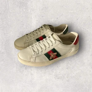 Gucci ace - Snygga vita sneakers från Gucci i skinn med klassiska gröna och röda ränder på sidan samt en broderad guldfärgad bi. Baksidan har detaljer i rött och grönt med textur. Skorna har snörning och platt sula, perfekt för dig som gillar lyxiga och trendiga märken.