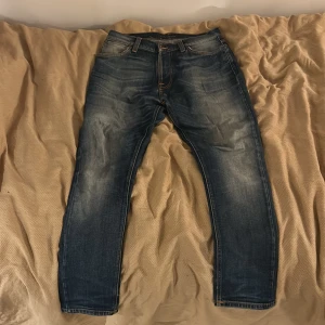 Blå jeans från Nudie Jeans Co - Säljer ett par klassiska blå jeans från Nudie Jeans Co med snyggt slitna detaljer och kontrastsömmar. Jeansen har normal passform, raka ben och fem fickor. Perfekta för dig som gillar en avslappnad men stilren look.