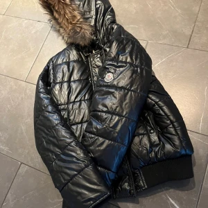 Svart Moncler pufferjacka med päls - Säljer en svart pufferjacka från Moncler med glansig yta och snygg päls på huvan. Jackan har dragkedja, snörning vid huvan och ribbade muddar. Perfekt för dig som vill ha en varm och stilren vinterjacka med exklusiv känsla.