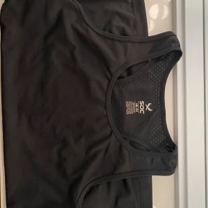 Svart linnetopp från SOC - Svart sportig linnetopp från SOC med meshdetaljer vid ryggen för extra ventilation. Toppen har en rundad hals och är ärmlös, perfekt för träning. Materialet är syntet och designen är stilren med logga framtill.