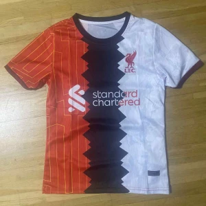 Liverpool FC fotbollströja röd/vit - Snygg Liverpool FC fotbollströja med unik design – ena halvan är röd med gula linjer, andra halvan är vit. Svart sicksackmönster i mitten och klubbmärke på bröstet. Kortärmad och tillverkad i lätt, andasvänligt material. Perfekt för dig som vill sticka ut på planen eller läktaren.