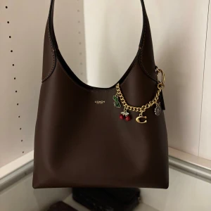 Brun brooklyn 28 väska från Coach  - Snygg mörkbrun axelväska från Coach. Väskan har minimalistisk design och diskret logga framtill. Perfekt storlek för att bära det viktigaste och passar till många outfits. Nypris 4100