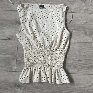 Prickig ärmlös topp från Gina Tricot - Supersöt ärmlös topp från Gina Tricot i cremevit med svarta prickar. Toppen har smockad midja och volangkant nertill. Tillverkad i ett lätt, strukturerat tyg som känns luftigt och skönt. Super fint skick aldrig använd🥰skriv privat för andra frågor 