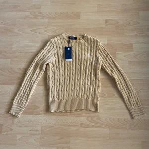 Beige stickad tröja Polo Ralph Lauren - Helt nu testad 1 gång. snygg beige kabelstickad tröja från Polo Ralph Lauren med klassisk rund hals och vit broderad logga på bröstet. Tröjan har långa ärmar och ribbade muddar vid ärmslut och nederkant. Perfekt för dig som gillar stilrena och tidlösa plagg.