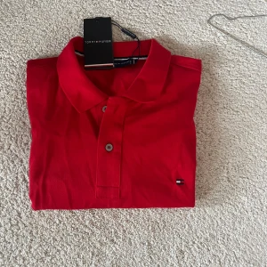 Röd pikétröja Tommy Hilfiger - Snygg röd pikétröja från Tommy Hilfiger med klassisk krage och två knappar framtill. Tröjan har korta ärmar och är tillverkad i 100% bomull. Diskret Hilfiger-logga broderad på bröstet. Perfekt för dig som gillar stilrena och sportiga plagg.