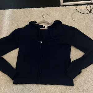 Svart hoodiejacka från Gina Tricot - Mörkblå hoodiejacka från Gina Tricot med dragkedja framtill och huva. Jackan har lång ärm och en enkel, stilren design som passar perfekt till vardags. Tillverkad i mjukt material för en bekväm känsla.