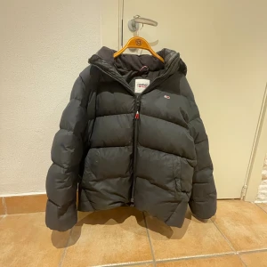 Svart pufferjacka från Tommy Jeans - Svart pufferjacka från Tommy Jeans med huva och dragkedja framtill. Jackan är vadderad och har en klassisk, boxig passform. Perfekt för kalla dagar och har en diskret logga på bröstet. Fodret är mjukt och jackan är riktigt skön att bära.