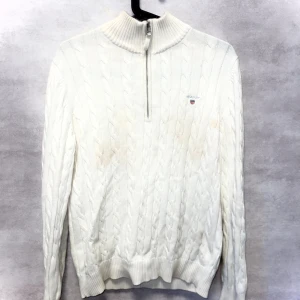 Vit kabelstickad half zip tröja GANT - Snygg vit kabelstickad tröja från GANT med halv dragkedja och hög krage. Tröjan har broderad logga på bröstet och är gjord i mjuk premium bomull. Perfekt för dig som gillar klassisk och clean stil med lite preppy vibe.