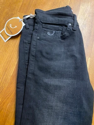 Svarta jeans från Jacob Cohen - Snygga svarta jeans från Jacob Cohen med klassisk femficksdesign och diskret J-broderi på myntfickan. Jeansen har raka ben och är tillverkade i ett mjukt denimtyg med stretch för extra komfort. Perfekta för en stilren och modern look.