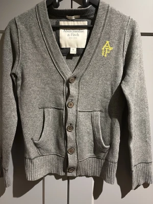Grå kofta från Abercrombie & Fitch - Grå kofta med knappar framtill från Abercrombie & Fitch. Koftan har två fickor, ribbade muddar och en gul broderad logga på bröstet. Djup V-ringning och mjukt stickat material gör den både snygg och bekväm. Skulle rekommendera storlek S-Xs