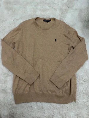 Beige stickad tröja Polo Ralph Lauren - Snygg beige stickad tröja från Polo Ralph Lauren i slim fit-modell. Tröjan har rund halsringning, långa ärmar och den klassiska broderade loggan på bröstet. Perfekt för lager-på-lager och enkel att matcha med olika outfits. Liten blå fläck på ryggen men inget som syns