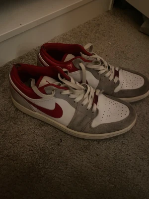 Nike Air Jordan 1 Low röd/vit/grå - Snygga Nike Air Jordan 1 Low sneakers i vitt, grått och rött. Skorna har klassisk röd swoosh, röd insida och broderad Air Jordan-logga på hälen. Ovandelen är i läder och mocka med perforerad tåbox och vita skosnören. Perfekta för dig som gillar streetwear.