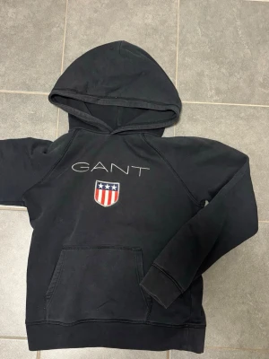 Svart hoodie från GANT med logga - Svart hoodie från GANT med broderad logga och sköld på bröstet. Klassisk känguruficka framtill och huva. Snygg och enkel design som passar perfekt till jeans. Mjuk bomullskänsla och bekväm passform. 