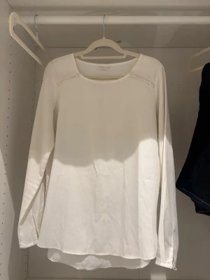 Vit långärmad blus med rund hals - Stilren vit blus med rund halsringning och diskreta detaljer vid axlarna. Långärmad modell i lätt och mjukt tyg som faller snyggt. Perfekt att matcha med jeans eller kjol för en clean look.