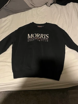 Svart sweatshirt från Morris - Svart sweatshirt från Morris med vit tryckt logga och texten 'Morris Sporting Club' på bröstet. Rund halsringning och ribbade muddar vid ärmslut och nederkant. Perfekt för en avslappnad och stilren look.