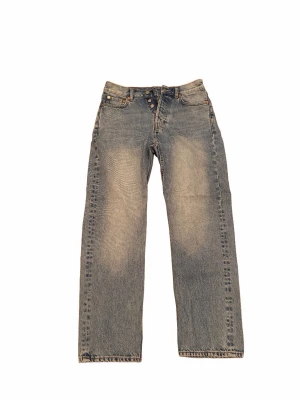 Blå raka jeans med knappgylf - Säljer ett par blå raka jeans med knappgylf och klassisk femficksmodell. Jeansen har en tvättad look med ljusare partier framtill och bakfickor. Materialet är kraftig denim i bomull och passformen är straight fit.