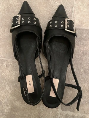 Svarta pumps med nitar från Nelly - Snygga svarta pumps från Nelly med spetsig tå, låg klack och bred rem med silverfärgade nitar och spänne. Skorna har tunna ankelband och är tillverkade i ett slätt material som ger en trendig look.