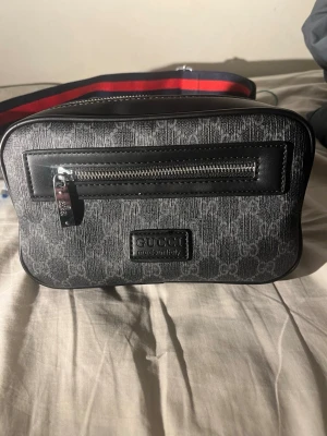 Svart axelväska från Gucci - Snygg svart axelväska från Gucci med klassiskt GG-mönster i grått. Väskan har en framficka med silverfärgad dragkedja och en bred axelrem i rött och blått. Tillverkad i slitstarkt canvasmaterial med detaljer i läder. 1.1!!! Använd några gånger säljs pågrund av inte använder 