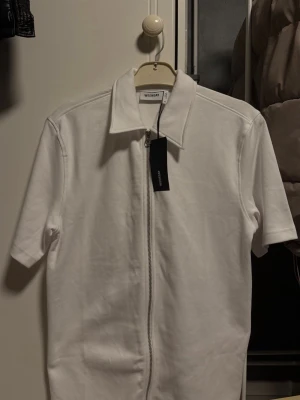 Vit kortärmad topp med dragkedja Weekday - Vit kortärmad topp från Weekday med krage och hel dragkedja framtill. Tillverkad i mjuk bomull och har en clean, minimalistisk look. Perfekt för dig som gillar stilrena plagg med en twist.